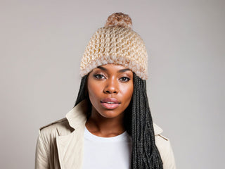 Woman with braids wearing a beige satin lined winter hat with a brown and white faux fur pom and trim.