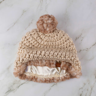 Beige satin lined winter hat with a brown and white faux fur pom and trim.