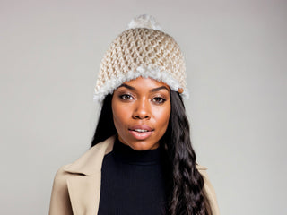 Woman wearing a beige satin lined winter hat with a white faux fur trim and pom.