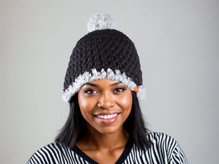 Woman wearing a black satin lined winter hat with a white faux fur pom and faux fur trim.