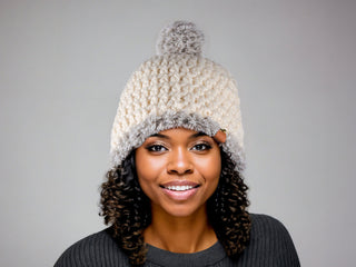 Woman wearing a cream satin lined winter hat with a gray faux fur trim and pom